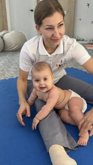 Kinetoterapeuta sprijina cu mainile un bebelus asezat pe o saltea albastra, in cabinet luminos; copilul zambeste si priveste spre camera.