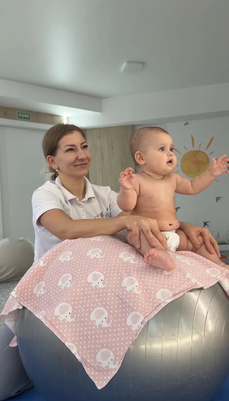 Kinetoterapeut KINETO TIME sprijină un bebeluș așezat pe o minge de gimnastică