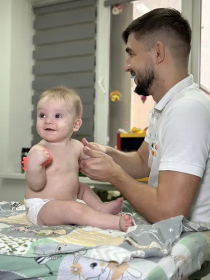 Bebeluș așezat pe o masă acoperită cu pătură cu model, ținând un obiect mic roșu, iar un adult zâmbește și îi susține brațul.