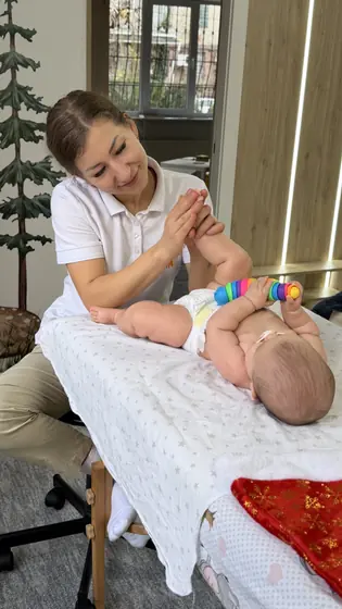 Femeie în tricou alb masează picioarele unui bebeluș întins pe o masă cu cearșaf alb; bebelușul ține o jucărie colorată.