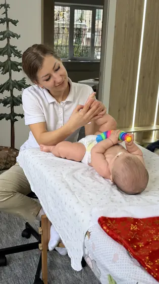 Specialistă în tricou alb ține cu ambele mâini piciorul unui bebeluș întins pe spate pe masa de masaj; bebelușul ține o jucărie colorată.