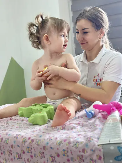 Copil așezat pe masa de masaj, susținut de un adult; jucării din burete și o coală tipărită pe cearșaf.