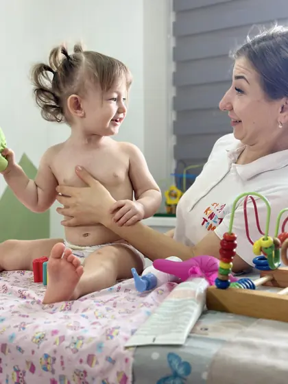Copil mic fără tricou, așezat pe masa de masaj, sprijinit cu mâna de o persoană adultă; amândoi zâmbesc, iar pe masă sunt jucării colorate din lemn.