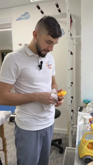 Barbat adult in picioare intr-un cabinet, privind in jos spre un obiect mic portocaliu din maini; in spate, perete alb decorat si scara de perete.