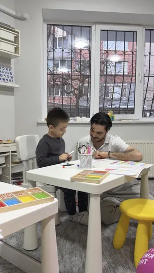 Un copil și un adult stau la o măsuță albă și lucrează cu foi colorate; în prim-plan sunt cuburi de lemn, iar în fundal o fereastră mare cu gratii.